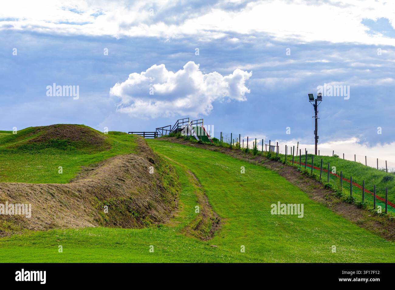 Grüne künstliche Röhrengleiter mit Treppen und Rampe steht auf einem grasbewachsenen Hügel. Schotterwege schlängeln sich über die Wiese unter einem dramatischen bewölkten Himmel. Zaun und Beleuchtung Stockfoto