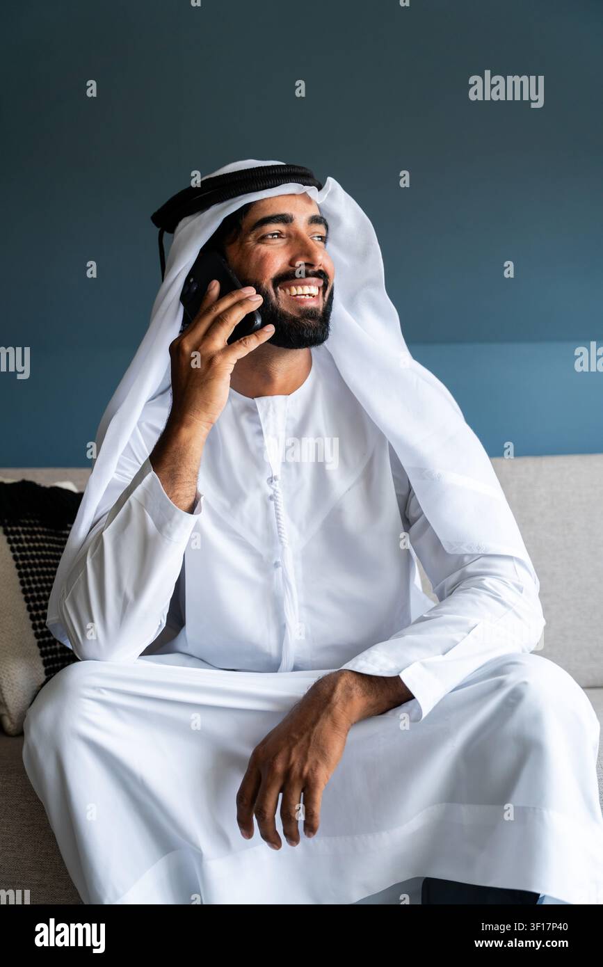 Emirati-Mann mit Bart, der traditionelle Kandura und Ghutra trägt, sitzt auf einem Sofa und telefoniert, drückt Glück und gute Kommunikation aus Stockfoto