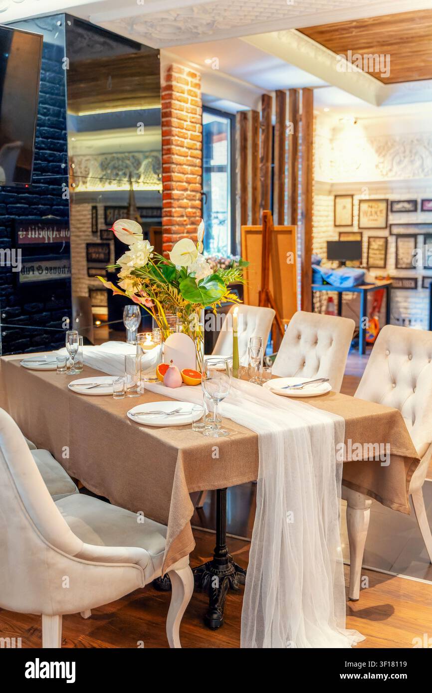 Ein Esstisch ist mit Tellern, Gläsern und Blumen arrangiert. Die Szenerie befindet sich in einem Restaurant am Tag mit einer warmen und einladenden Atmosphäre. Stuhl Stockfoto