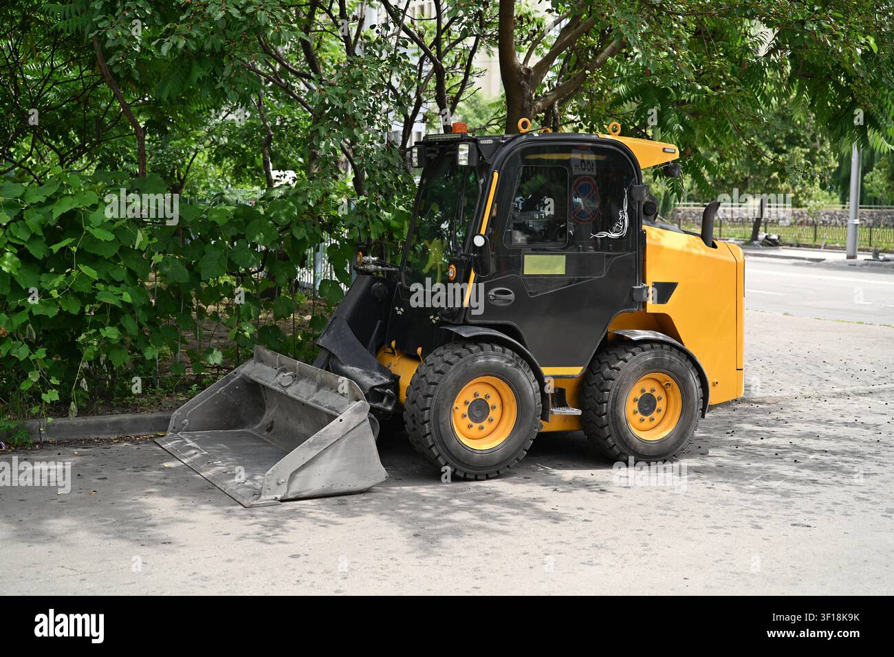Gelber Skid-Steer-Lader, der auf einer asphaltierten Straße in der Nähe von grünen Bäumen für die Stadtpflege geparkt ist. Stockfoto