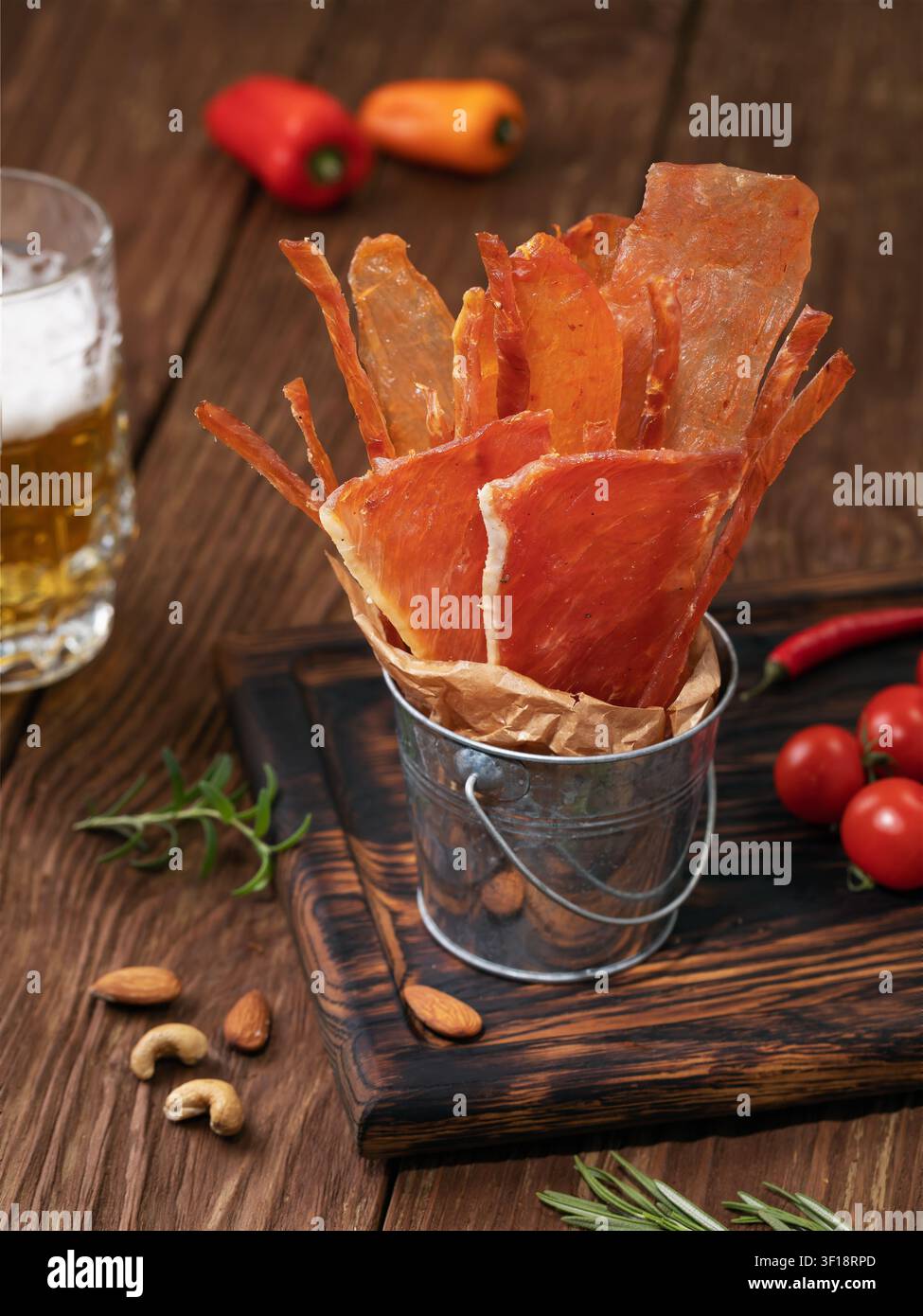 Scheiben und Streifen von getrocknetem geräuchertem Fleisch in einem Metallkübel mit einem Glas Bier, Nüssen und Gewürzen auf einem Holztisch Stockfoto