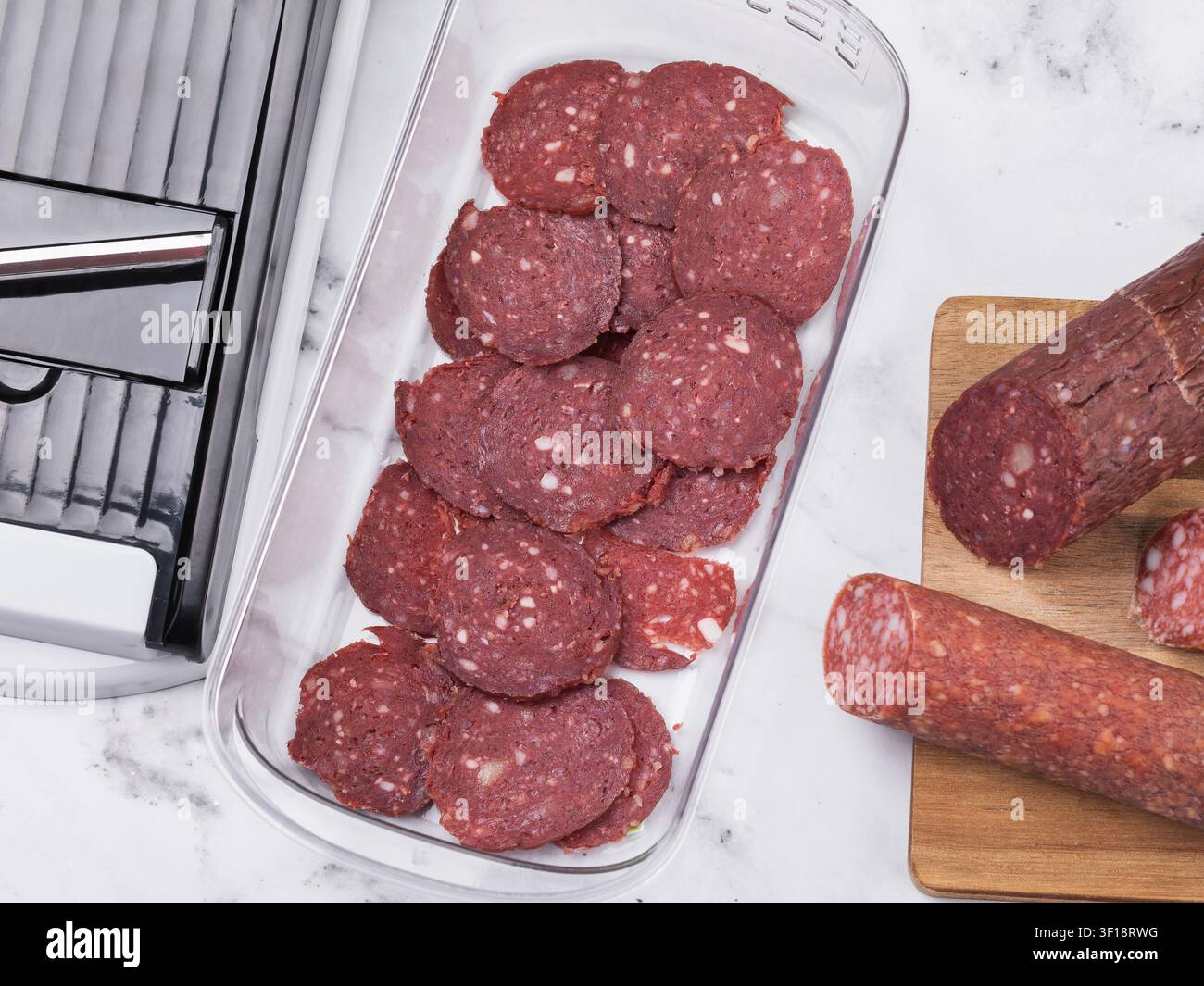 Geschnittene getrocknete Salami-Rundstücke in einem Behälter mit Schneidewerkzeug auf Marmortisch, Ansicht von oben Stockfoto