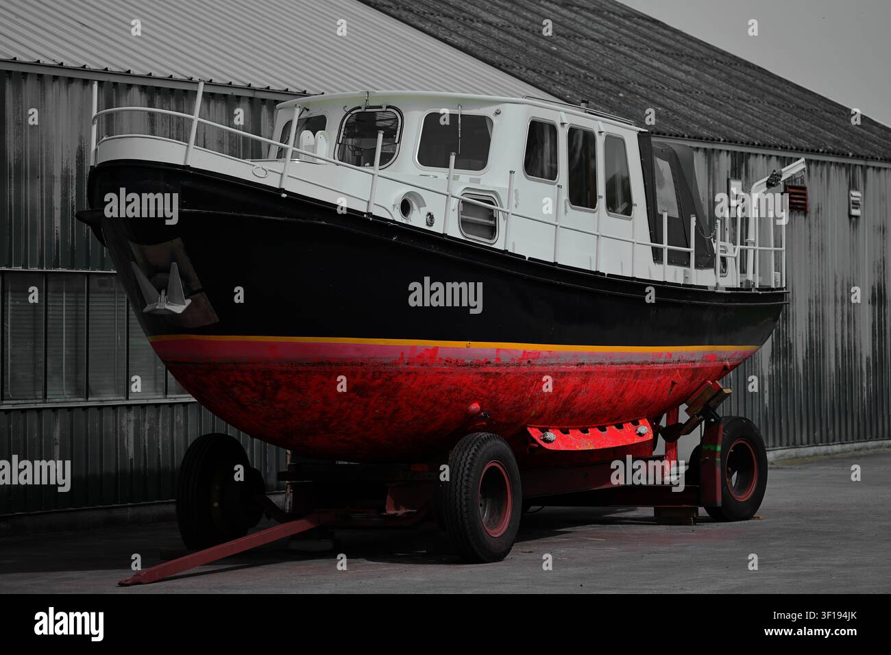 Ein verwittertes weiß-schwarzes Kabinenboot ruht auf einem Radanhänger neben einem wellblechverkleideten Schuppen in Zuid Holland, Niederlande, wobei sein roter Rumpf sichtbar ist. Stockfoto