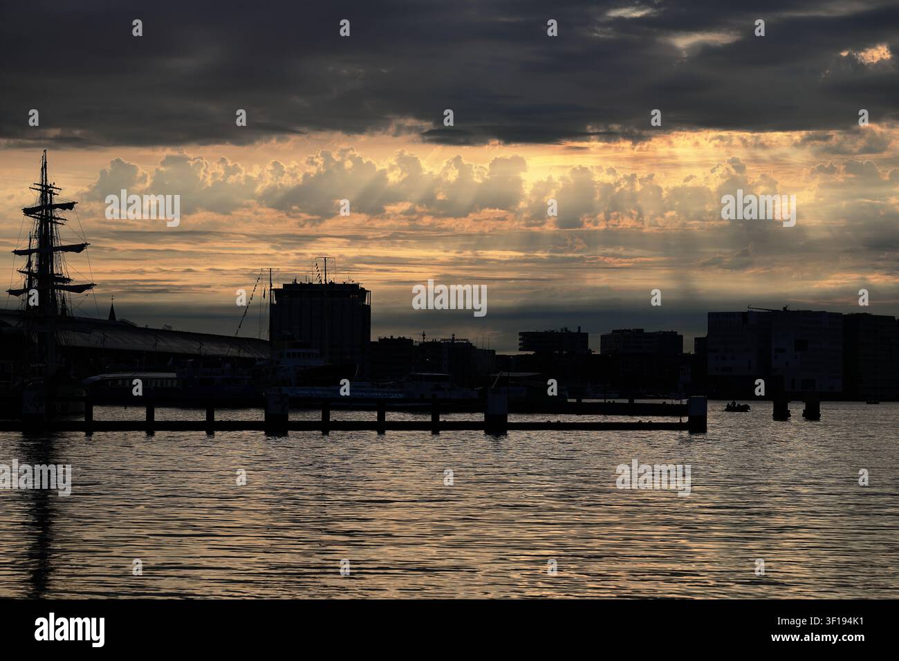 Die Skyline von Amsterdam am Wasser bildet eine dunkle Silhouette unter dichten, geschichteten Wolken, mit einem hohen Segelschiffmast und welligem Wasser im Vordergrund. Stockfoto