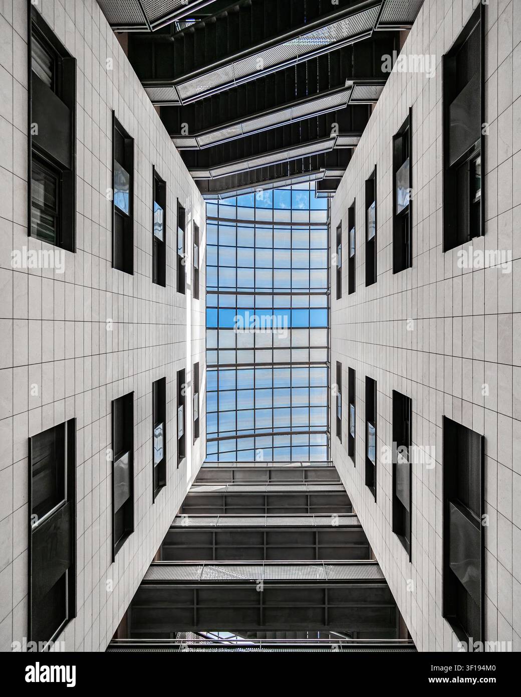 Aufwärts gerichtete architektonische Ansicht durch ein schmales Atrium unter einem Glasdach, eingerahmt von weißen Betonwänden, scharfen geometrischen Winkeln, klaren Linien, stark Stockfoto
