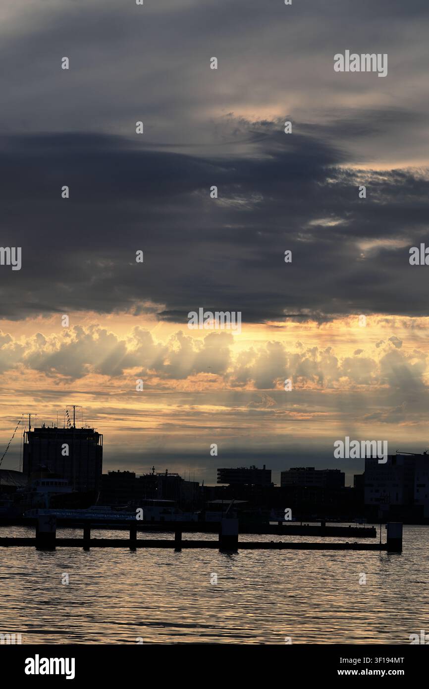Die dunkle Skyline von Amsterdam liegt unter einer Lücke in geschichteten Wolken, während niedrige Sonnenstrahlen einen leuchtenden Streifen am Horizont über dem welligen Uferwasser beleuchten. Stockfoto