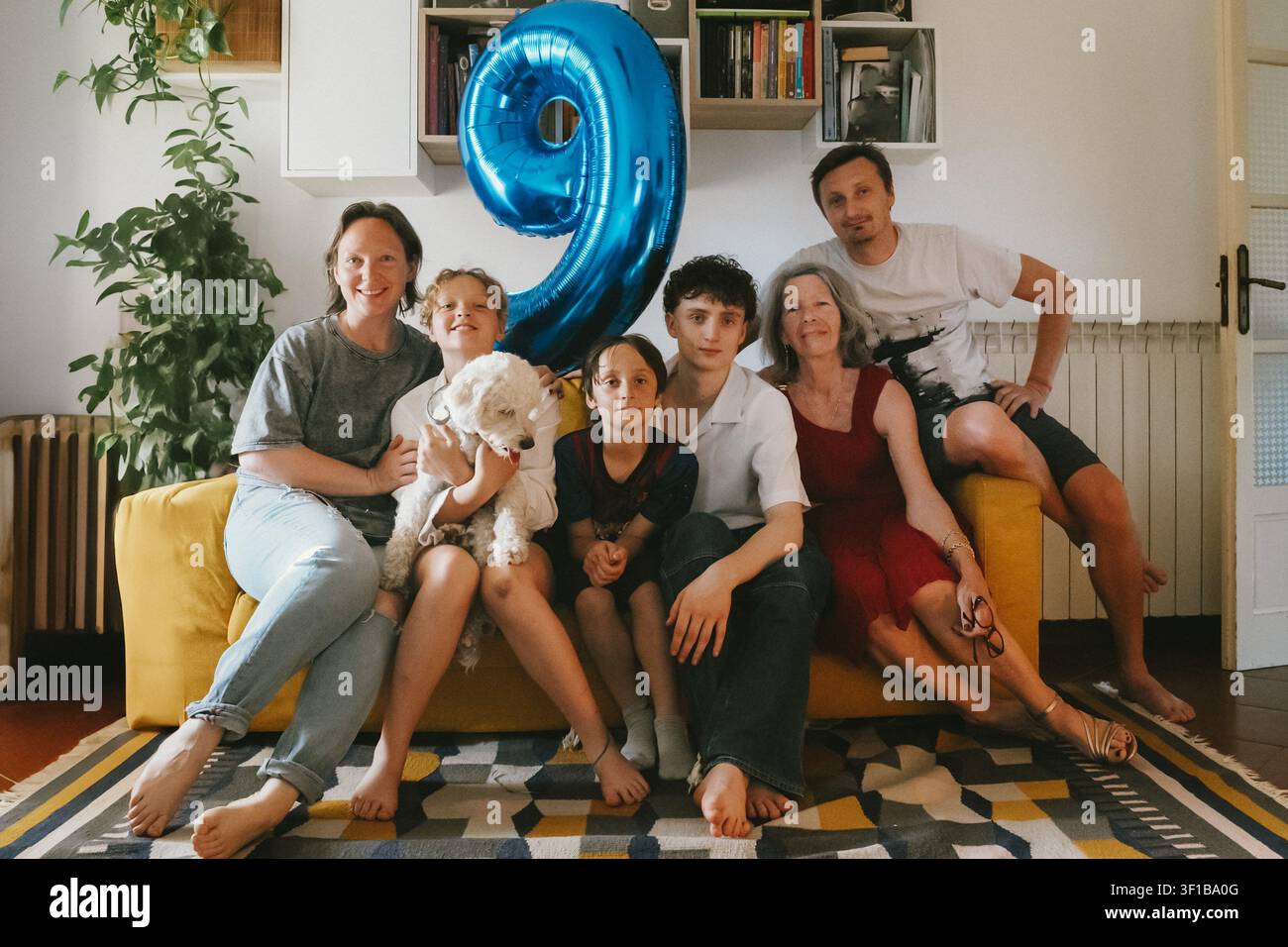 Fröhliche multigenerationale Familie, die sich während der Geburtstagsfeier eines Jungen zu Hause auf einem Sofa versammelt hat, mit einem großen blauen Luftballon mit der Zahl neun und einem Haustierhund. Stockfoto