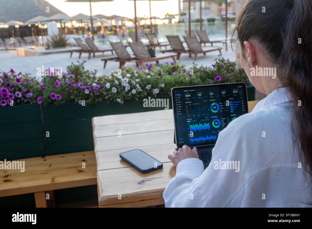 Frau in weißem Hemd sitzt an einem Holztisch und überprüft Finanzdaten und Diagramme auf einem Tablet-Bildschirm auf einer Außenterrasse eines Resorts. Stockfoto