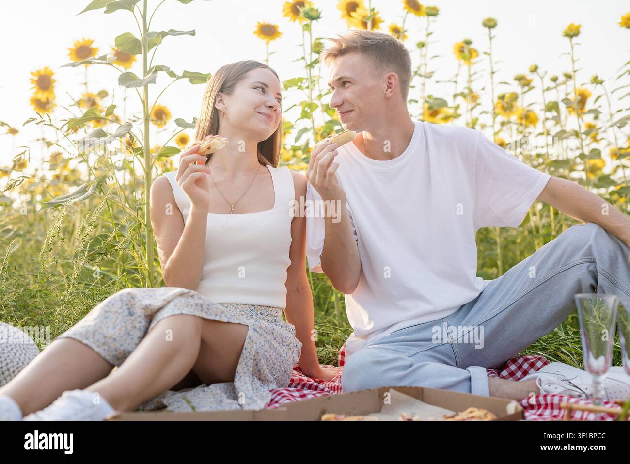 Ein junges Teenager-Paar in weißen Hemden, das bei Sonnenuntergang auf dem Sonnenblumenfeld picknickt. Pizza essen und Champagner trinken Stockfoto