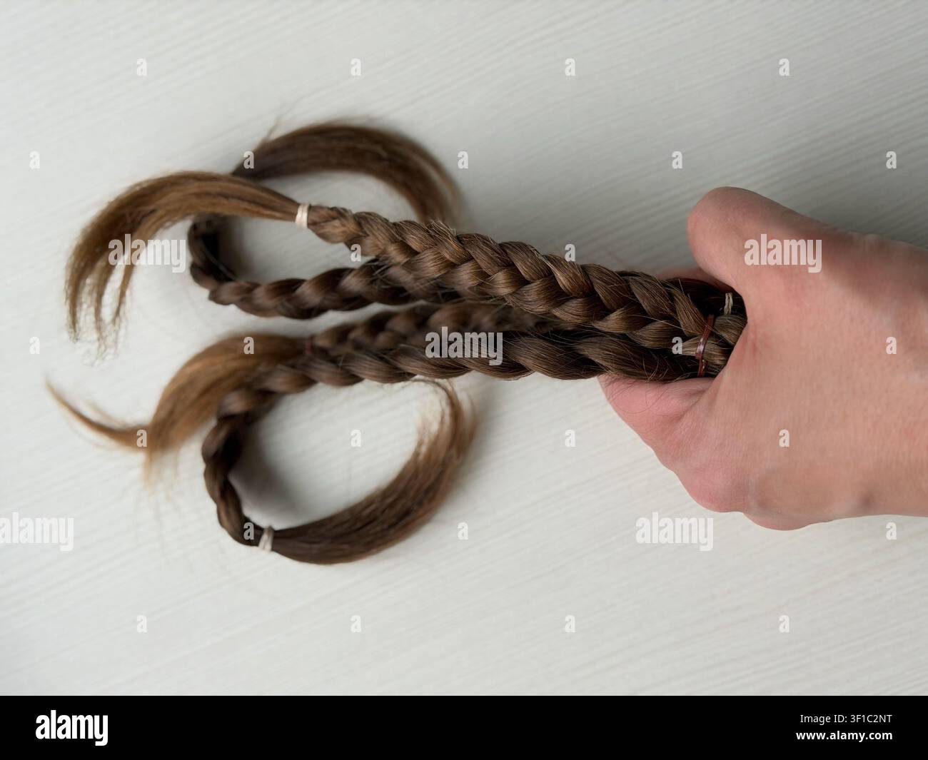 Menschliche Hand, die geschnittene geflochtene braune Haarsträhnen für eine Spende hält. Eine Nahaufnahme einer Person, die frisch geschnittene Zöpfe auf einem hellen Holzgrund hält. Stockfoto