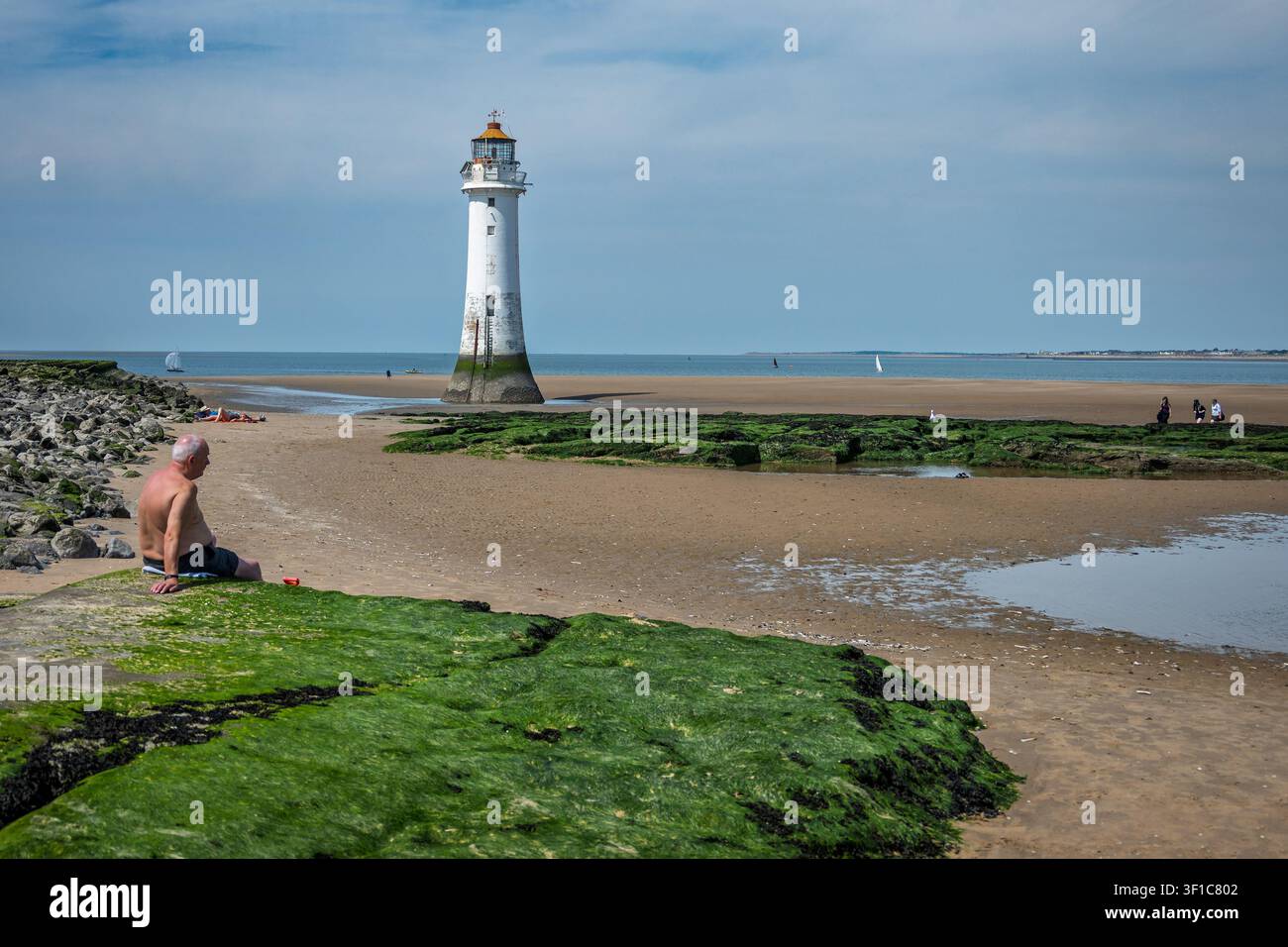 Ein Mann sitzt auf einem moosigen Felsen und genießt den sonnigen Tag am sandigen Pool in New Brighton. Der Leuchtturm ragt im Hintergrund empor, während sanfte Wellen Stockfoto