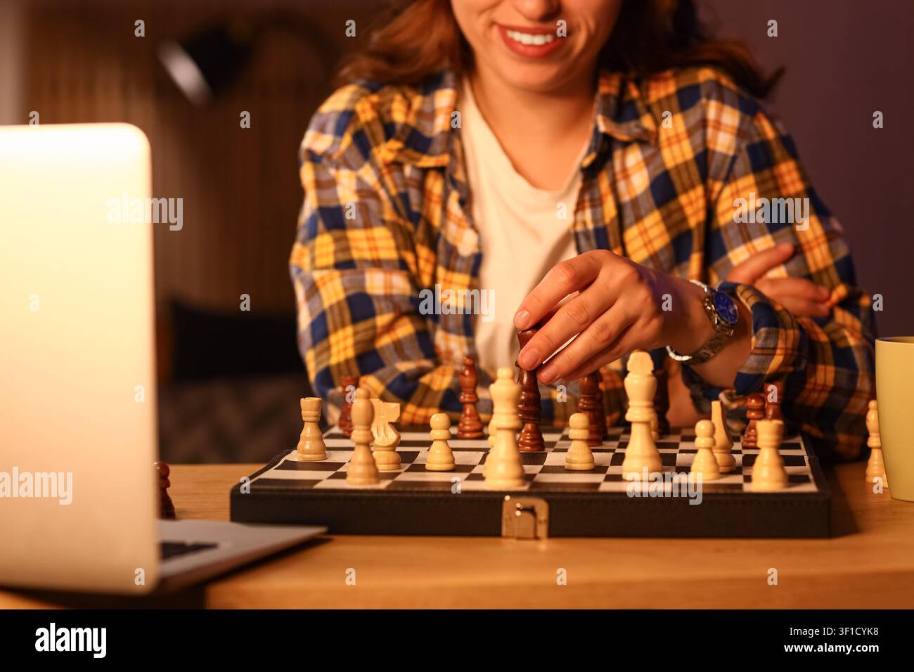 Lächelnde Frau spielt Schach an einem Holztisch mit einem Laptop in der Nähe, Strategie und intellektuelle Herausforderung Stockfoto