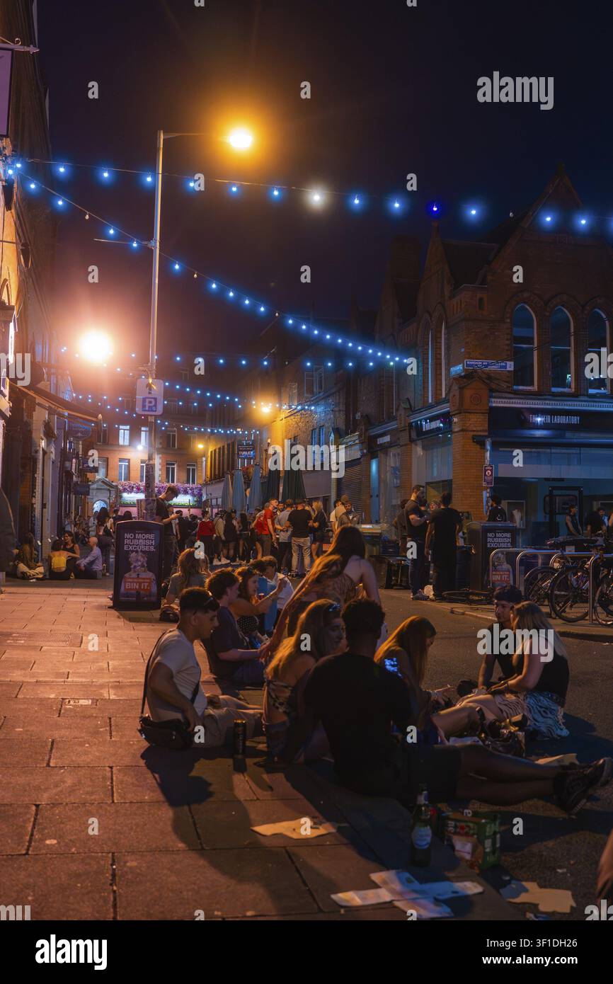 Gruppe von Menschen, die auf einer Straße sitzen, umgeben von warmen Lichtern und Laternen, Nachtleben, Dublin, Irland Stockfoto