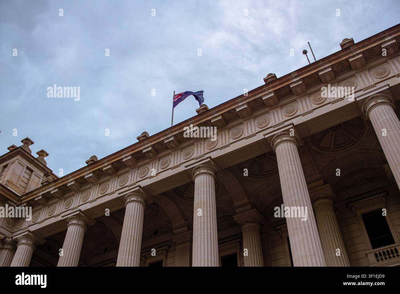 Ein majestätisches neoklassizistisches Gebäude mit hohen Steinsäulen und der australischen Nationalflagge, die sich gegen einen düsteren, bewölkten Himmel weht. Stockfoto