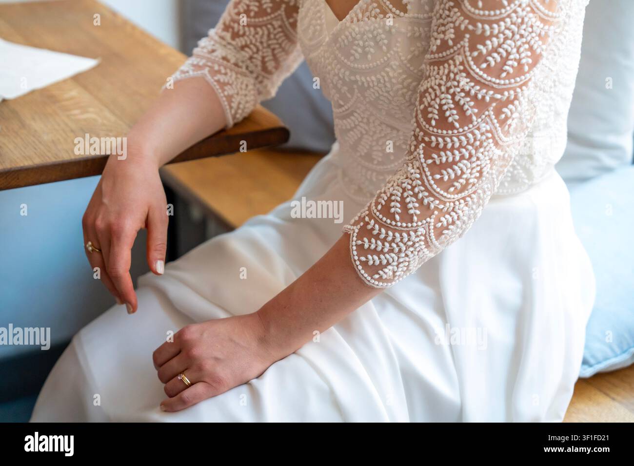 Braut in weißem Hochzeitskleid mit Spitzenärmeln, die auf einer Holzbank sitzt, Nahaufnahme von Händen und Ringen Stockfoto