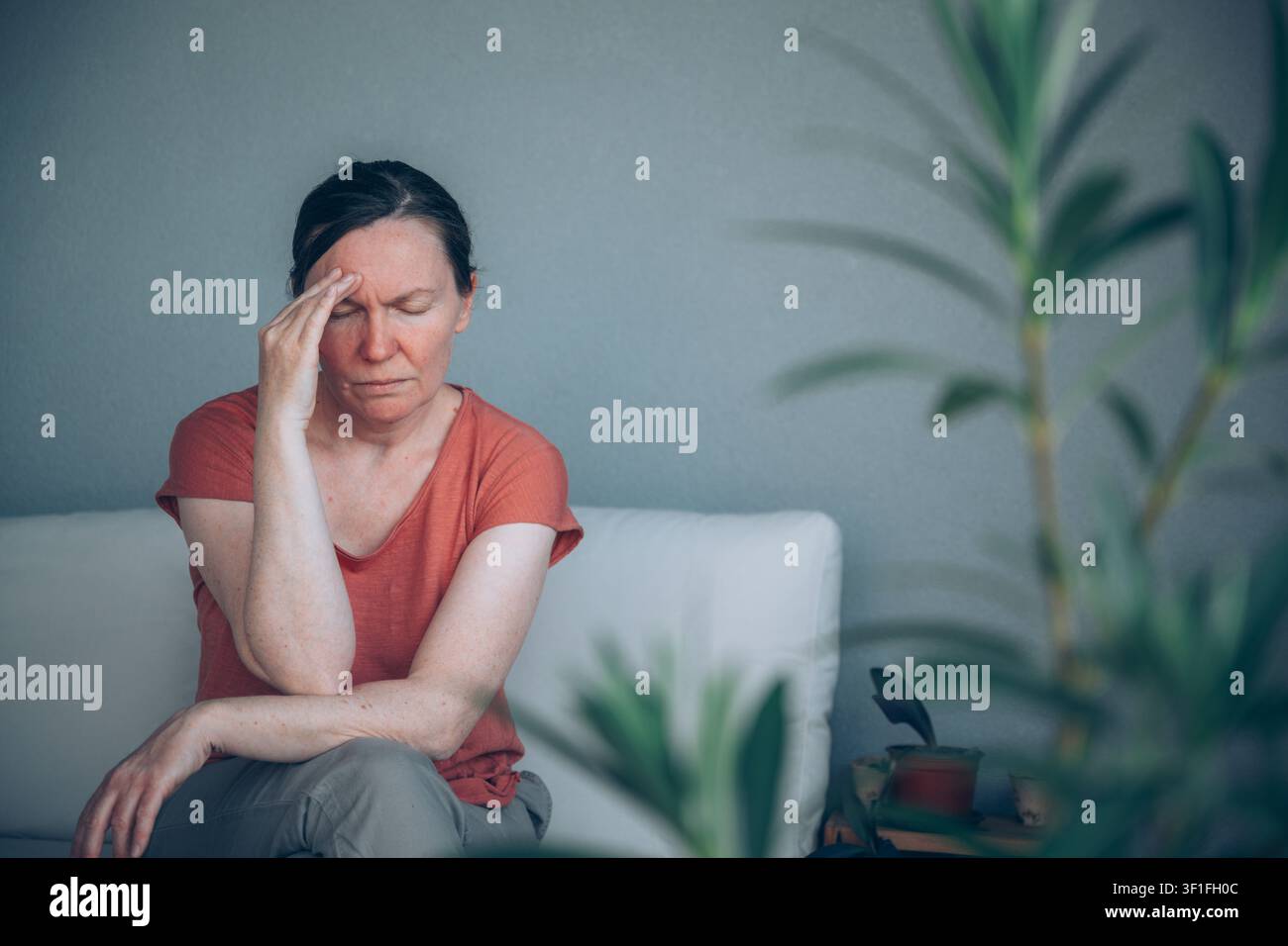 Reife Frau leidet unter starken Kopfschmerzen, während sie auf dem Balkonsofa sitzt, gesehen durch Oleanderblätter. Migräne-, Stress- und Gesundheitsproblemkonzept. Stockfoto