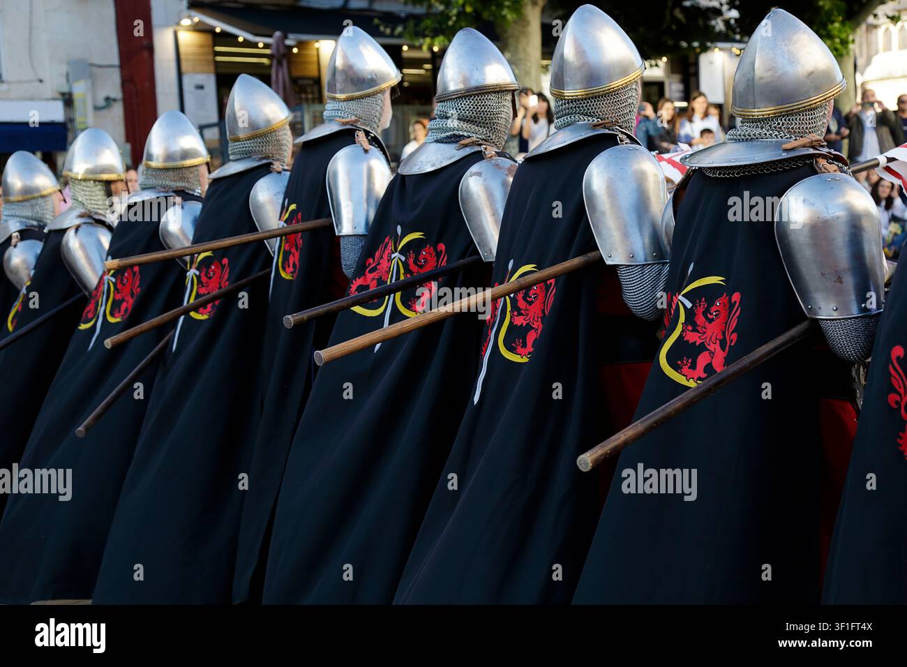 Eine Reihe von mittelalterlichen Reenactern, die Stahlhelme, Kettenhemden und schwarze Umhänge mit einem roten Löwenemblem tragen und lange Stangenwaffen bei einem Outdoor-P Stockfoto