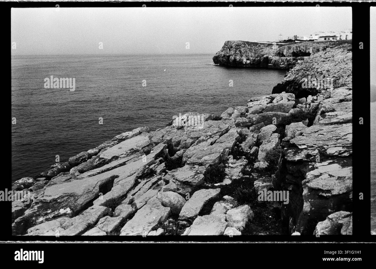Gebrochene Steinregale führen über eine raue Küste zu ruhigem Wasser und einer fernen Klippenansiedlung. Stockfoto