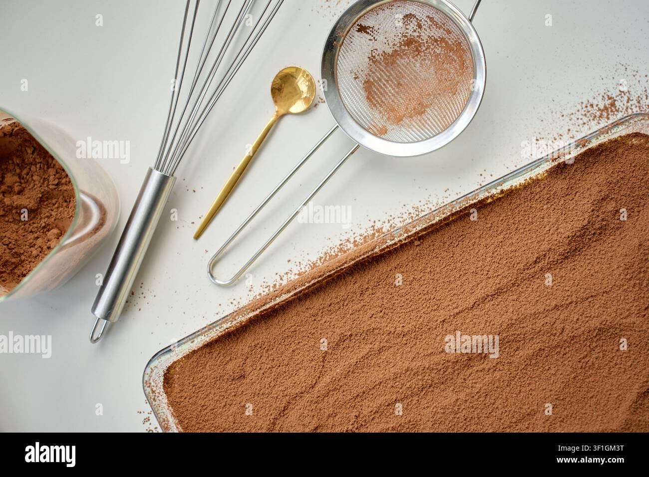 Tiramisu-Dessert mit Kakaopulver bedeckt in der Backform, mit Sieb und Kakaoschüssel auf dem Tisch. Konzept für hausgemachtes Dessert und italienische Süßspeise Stockfoto