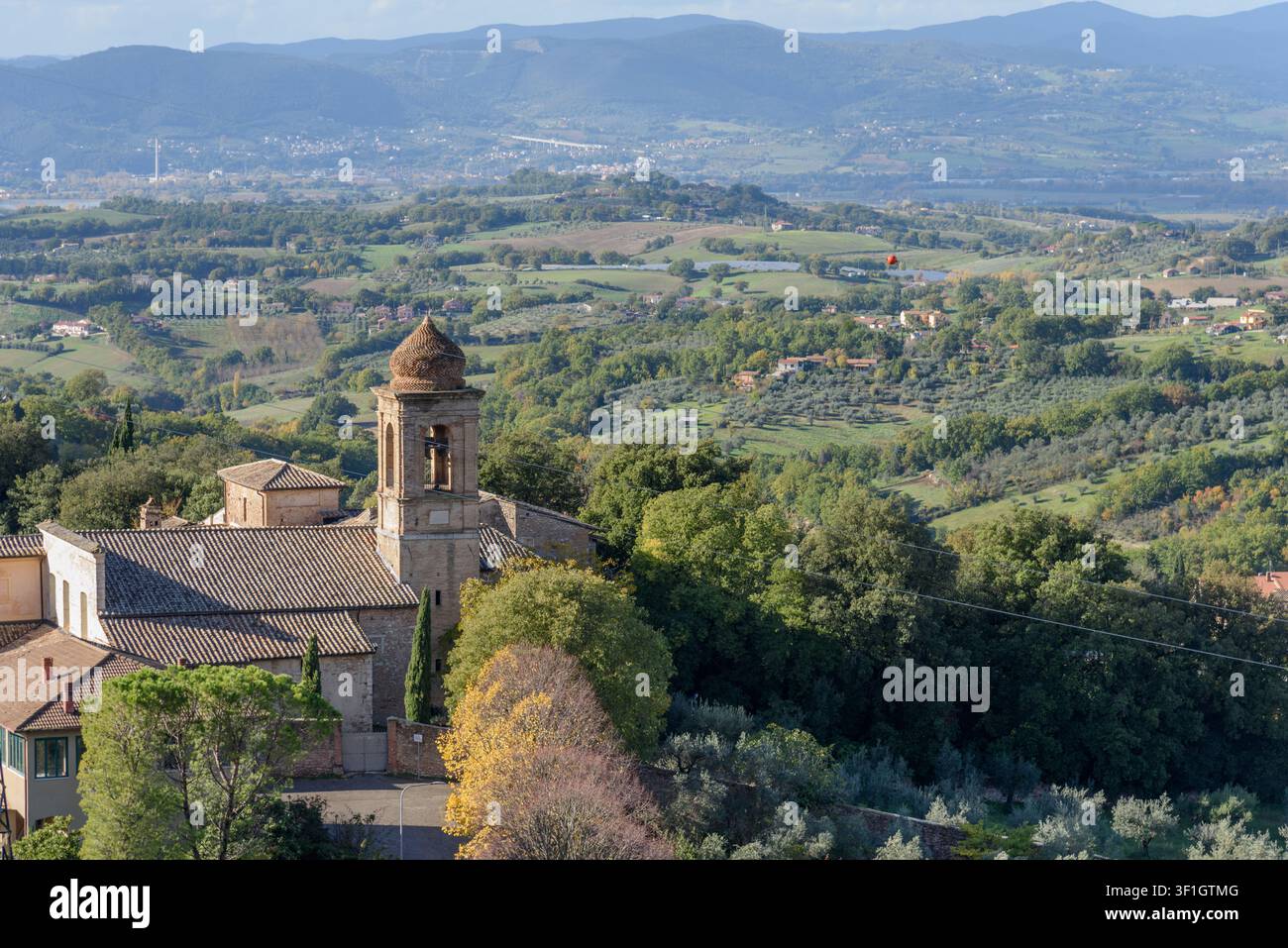 Panoramische Hochwinkelaufnahme einer mittelalterlichen Kirche mit einem einzigartigen Glockenturm, der über sanfte Hügel, Olivenhaine und Berge in der Landschaft von U hinwegragt Stockfoto