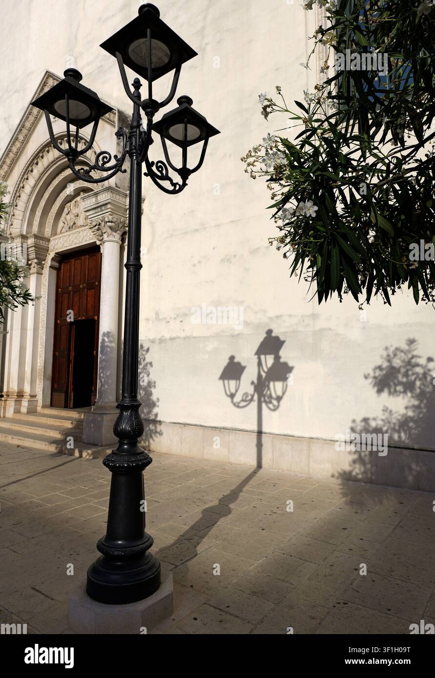 Verzierter schwarzer Straßenlaterne steht vor einer weißen Kirchenwand mit einem geschnitzten Torbogen, wirft einen langen Schatten. Italien, Apulien, Molfetta, 21. Juli Stockfoto