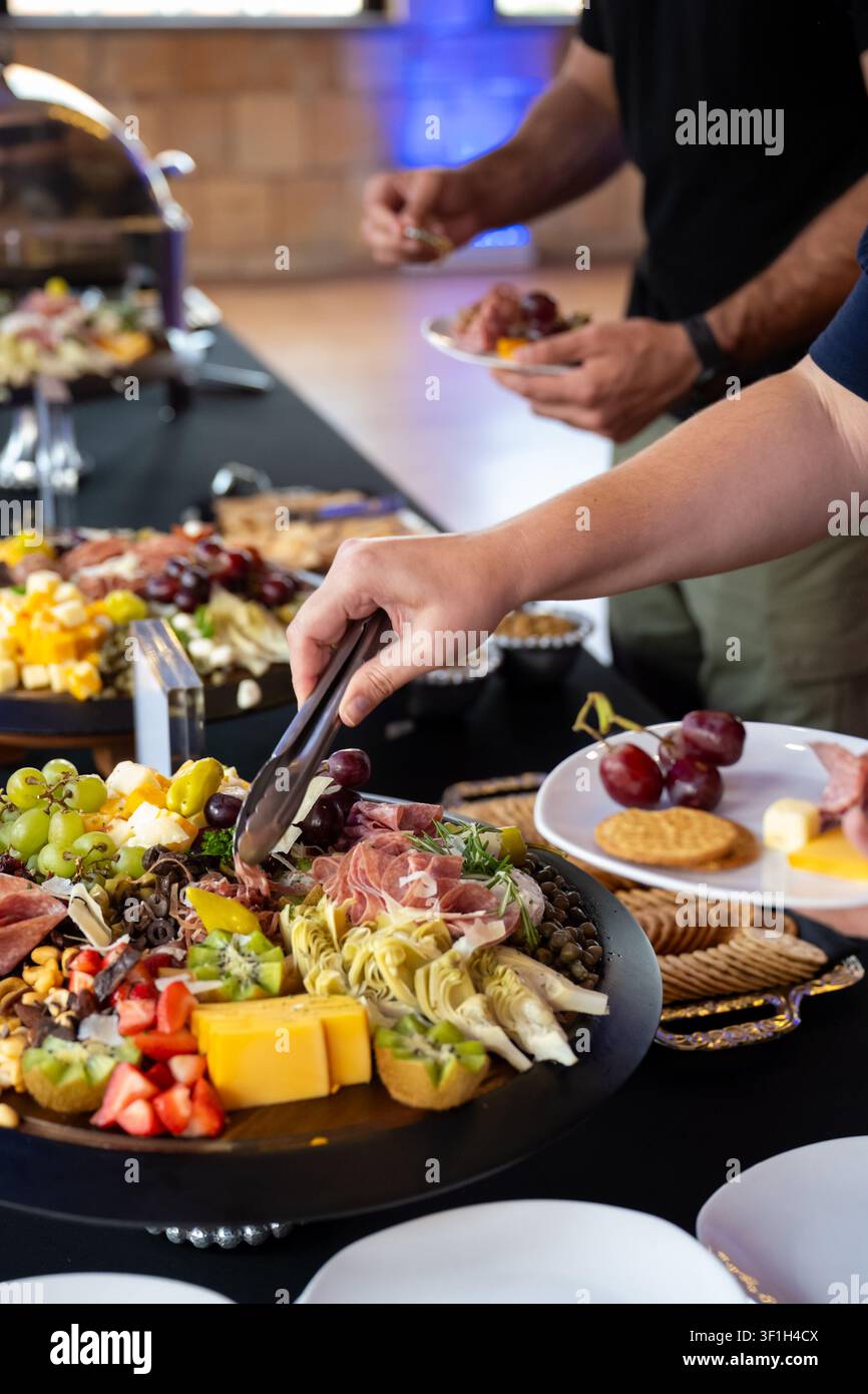 Gäste verwenden Servierzangen, um Käse, Obst, gepökeltes Fleisch, Cracker und Gemüse von einem Charcuterie-Tisch bei einer Indoor-Veranstaltung auszuwählen. Stockfoto