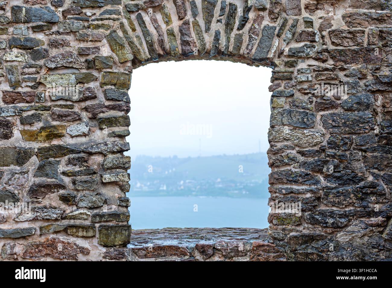 Alter Steinbogen, der einen nebligen Seeblick einrahmt. Verwitterte Steinmauer mit einer bogenförmigen Öffnung, die einen ruhigen See und eine entfernte neblige Uferlinie einrahmt Stockfoto