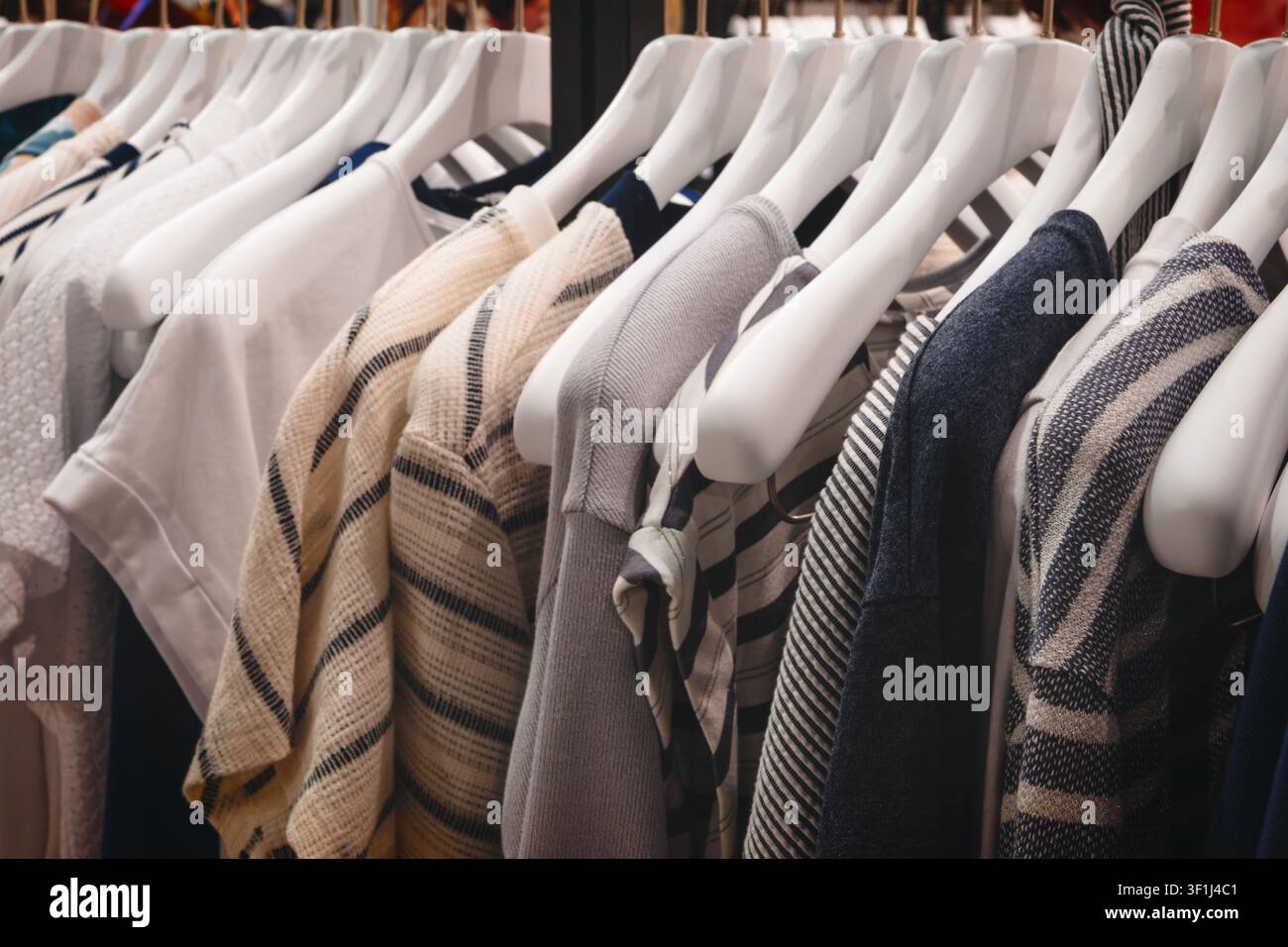 Weiße Plastikbügel halten zahlreiche Kleidungsstücke in einer Ladenauslage. Die Kleidungsstücke umfassen gestreifte, gestrickte und einfarbige Teile. Die Stoffe wirken weich, Stockfoto