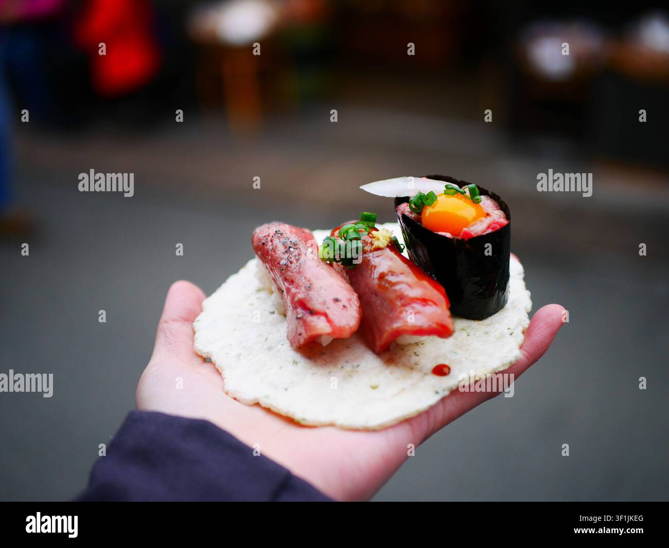 Eine Hand hält ein Stück Hida-Wagyu-Rindfleisch-Nigiri auf einem Reiscracker, das an einem japanischen Straßenfood-Markt an einem Stand gekauft wurde, mit weiteren Lebensmitteln und dem verschwommenen Markt-Hintergrund im Hintergrund. Stockfoto