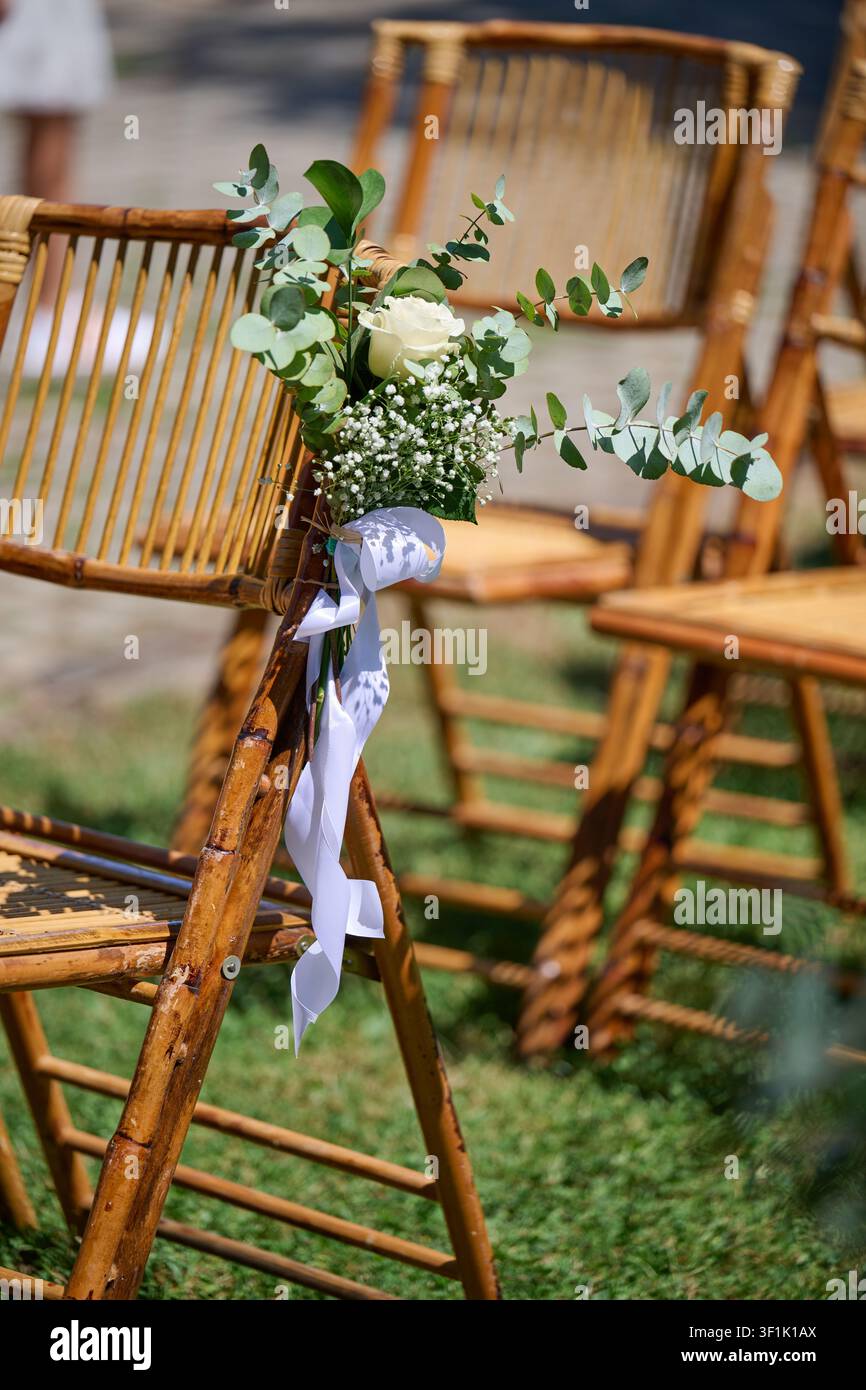 Rattan-Hochstuhl dekoriert mit einem eleganten Strauß aus einer weißen Rose, Schleierkraut und Eukalyptus, gebunden mit einem weißen Band, draußen aufgestellt für eine Stockfoto