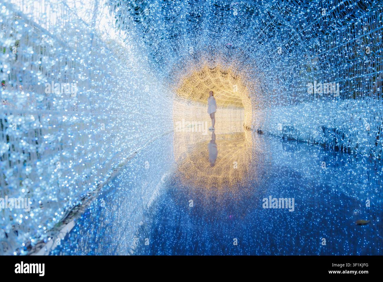 Eine Frau steht in einem faszinierenden immersiven Lichtinstallationstunnel mit brillanter blauer und goldener Beleuchtung, die sich auf einem polierten Boden spiegelt. Stockfoto