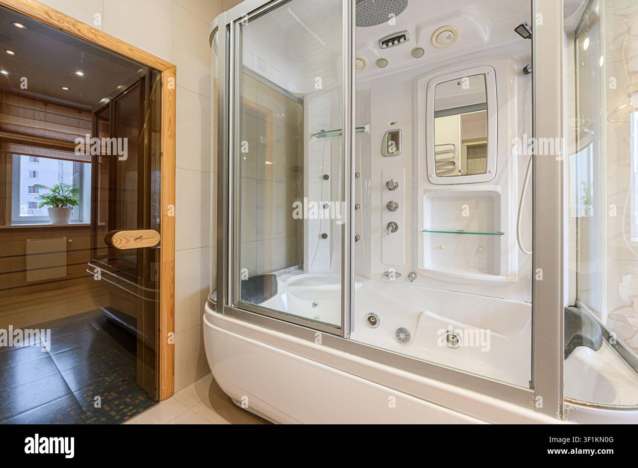 Ordentliches, beigefliestes Badezimmer mit weißem Waschbecken, einem Spiegel, einem Fenster mit einer Pflanze und einer Toilette. Die Gesamtästhetik ist sauber und gut beleuchtet Stockfoto
