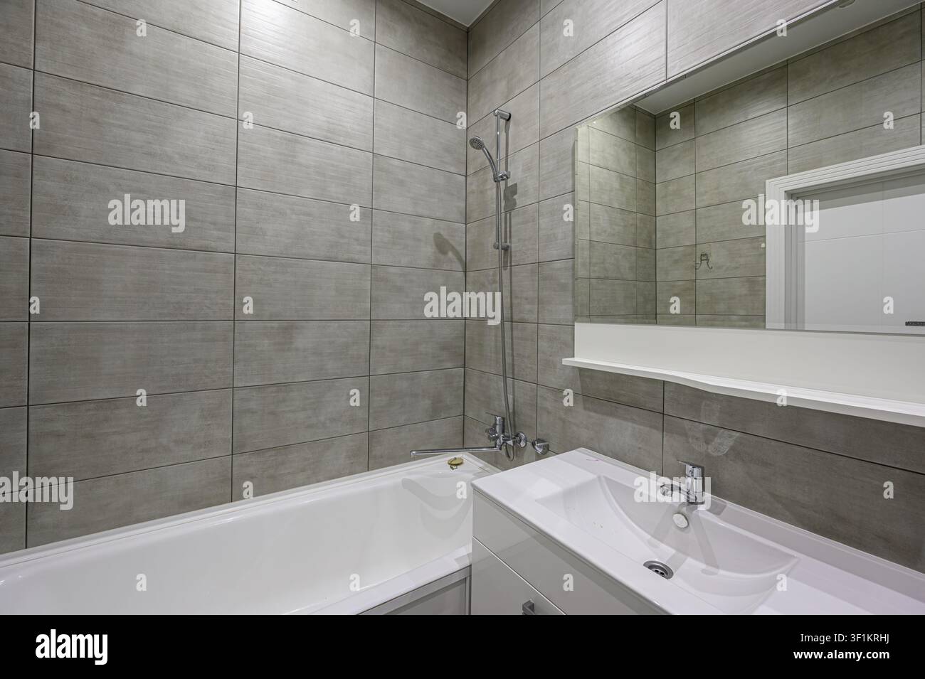 modernes Badezimmer mit grauen Fliesenwänden, einer weißen Badewanne mit Dusche und einem schlanken weißen Waschtisch mit einem großen Spiegel darüber Stockfoto
