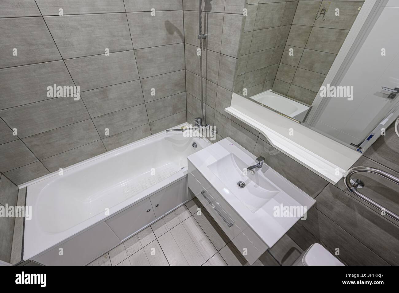 Modernes Badezimmer mit grauen Fliesenwänden, einer weißen Badewanne mit Dusche und einem schlanken weißen Waschtisch mit einem großen Spiegel darüber. Der Boden ist ebenfalls hell Stockfoto