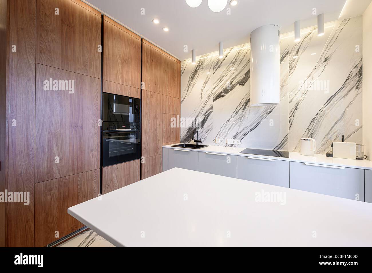 moderne Küche mit holzgetönten Schränken, einem schwarzen Backofen, grauen Arbeitsplatten, einer Marmor-Arbeitsrückwand und einer weißen Insel Stockfoto