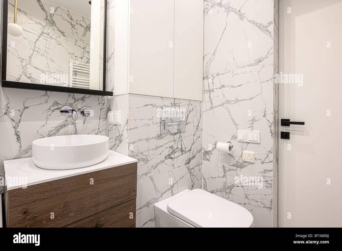 modernes Badezimmer mit weißer marmorierter Wandverkleidung, einem runden weißen Waschbecken auf einem Holzkabinett, einem schwarz gerahmten Spiegel und einer weißen Toilette Stockfoto
