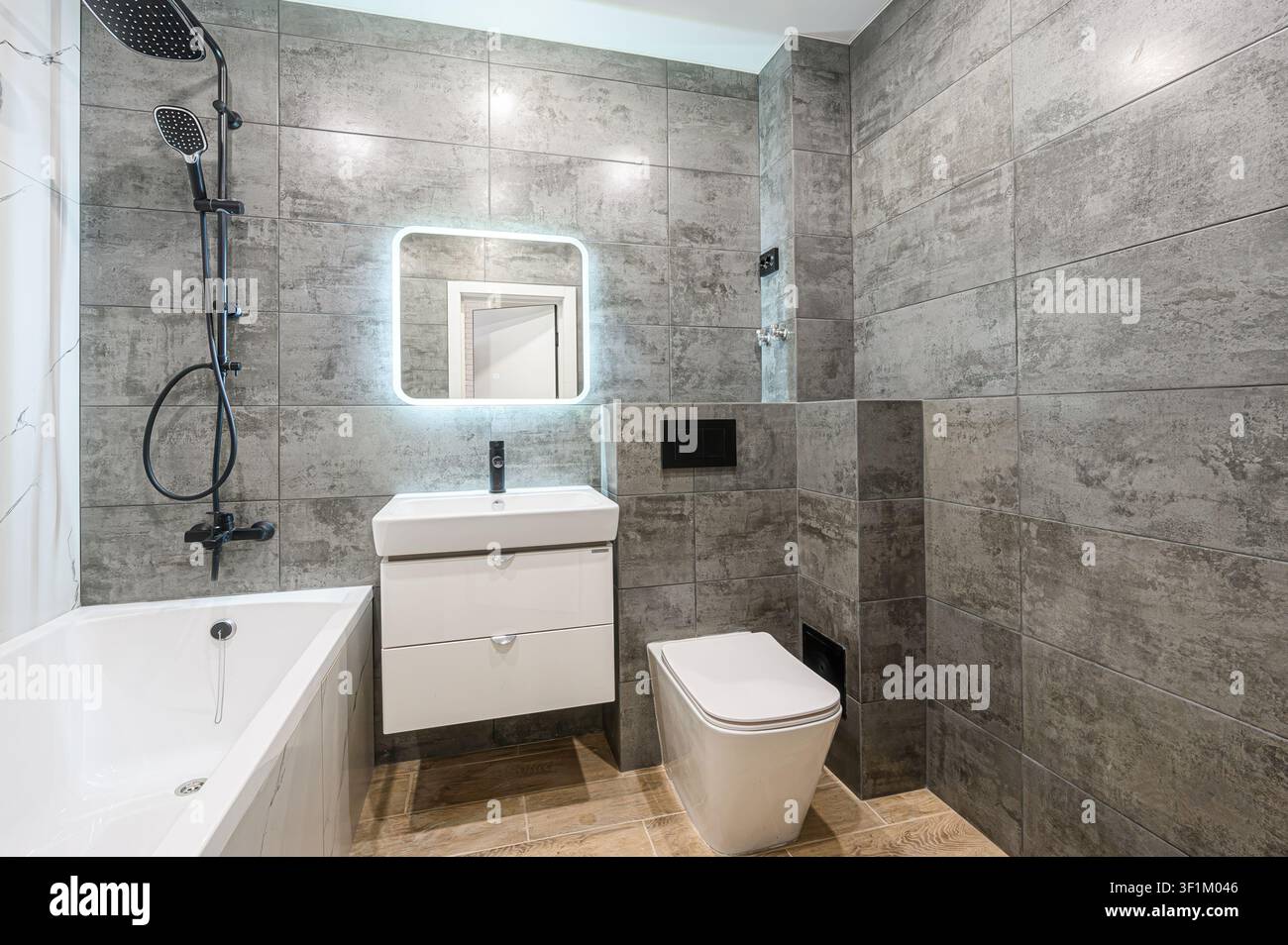 Modernes Badezimmer mit grauen Fliesenwänden, einem weißen Waschtisch, Toilette und Badewanne. Ein hinterleuchteter Spiegel verleiht einen Hauch zeitgenössischen Stils Stockfoto