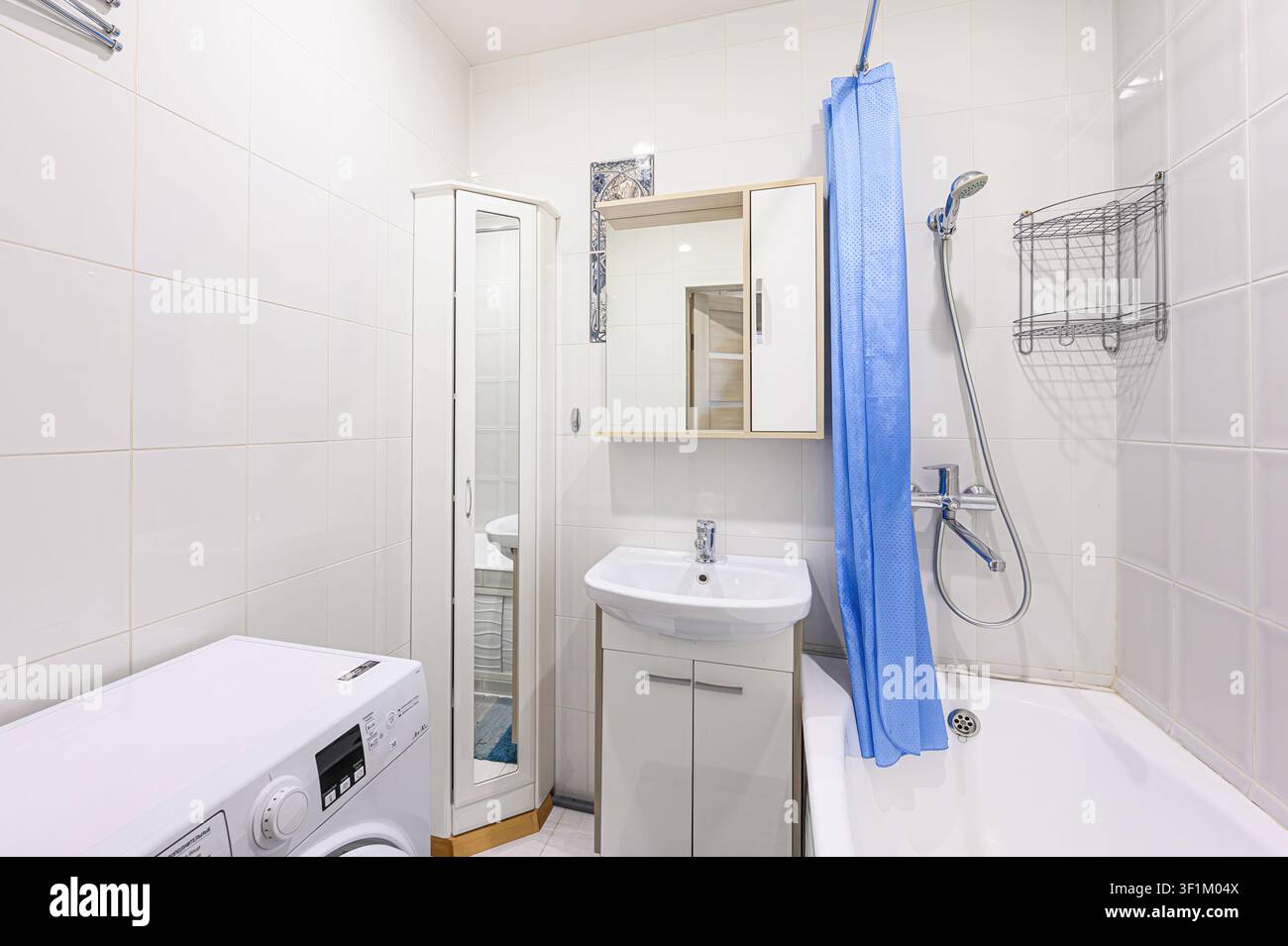 helles Badezimmer mit weiß gefliesten Wänden, ausgestattet mit einer Waschmaschine, Waschbecken mit Waschtisch, Spiegelschrank, Badewanne mit blauem Duschvorhang und einer Koralle Stockfoto