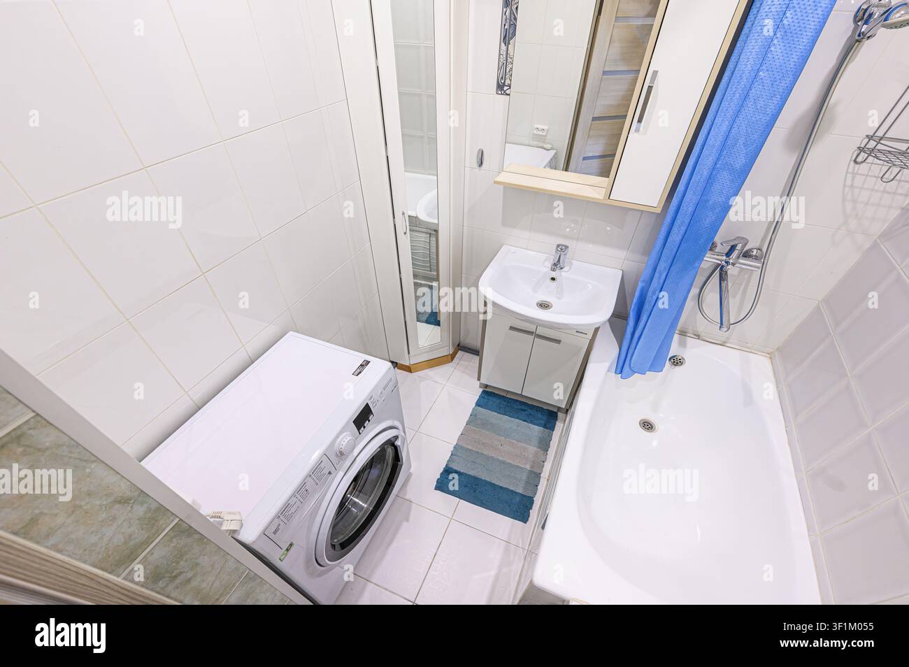 helles, kompaktes Badezimmer mit weißen Fliesen, einer Waschmaschine, einem Waschbecken, einer Badewanne mit blauem Vorhang und einem Spiegelschrank Stockfoto