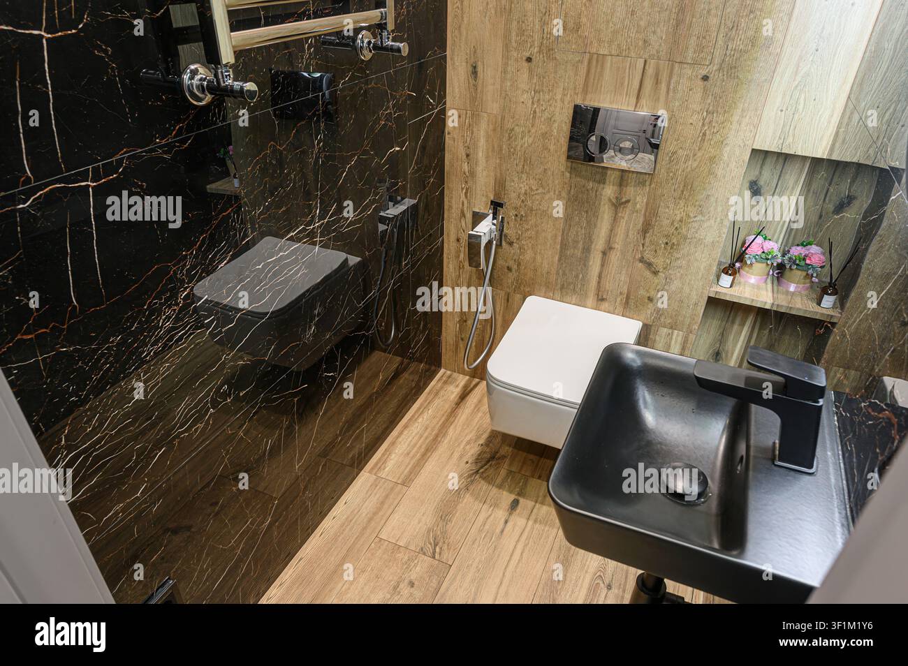 Stilvolles Badezimmer mit schwarzen Marmorwänden, hellen HolzAkzenten, modernen Armaturen einschließlich eines schwarzen Waschtischs, einer Toilette und einem beheizten Handtuchhalter Stockfoto