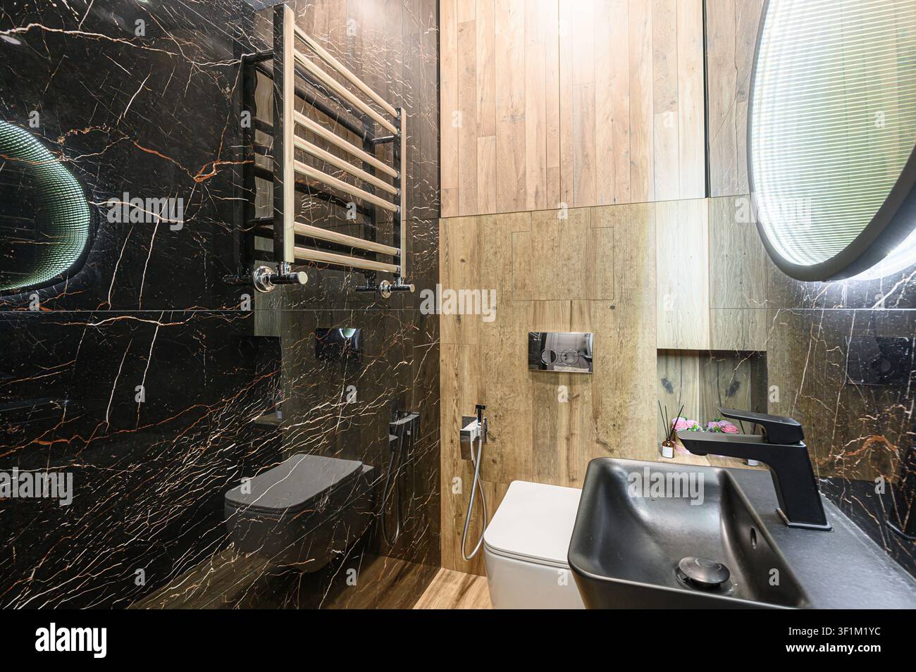 Stilvolles Badezimmer mit schwarzen Marmorwänden, hellen HolzAkzenten, modernen Armaturen einschließlich eines schwarzen Waschtischs, einer Toilette und einem beheizten Handtuchhalter Stockfoto