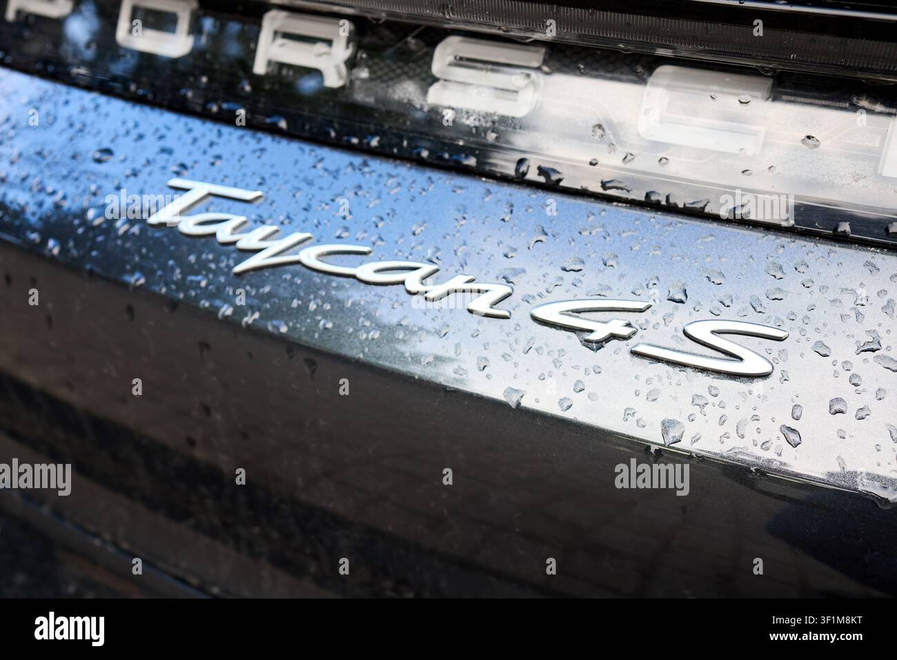 Berlin, Deutschland - 10. Juli 2026: Nahaufnahme des Chrom-Emblems und der Lichtleiste des Porsche Taycan 4S auf einem nassen dunklen Metallkarosserie mit Regentropfen, Sch Stockfoto