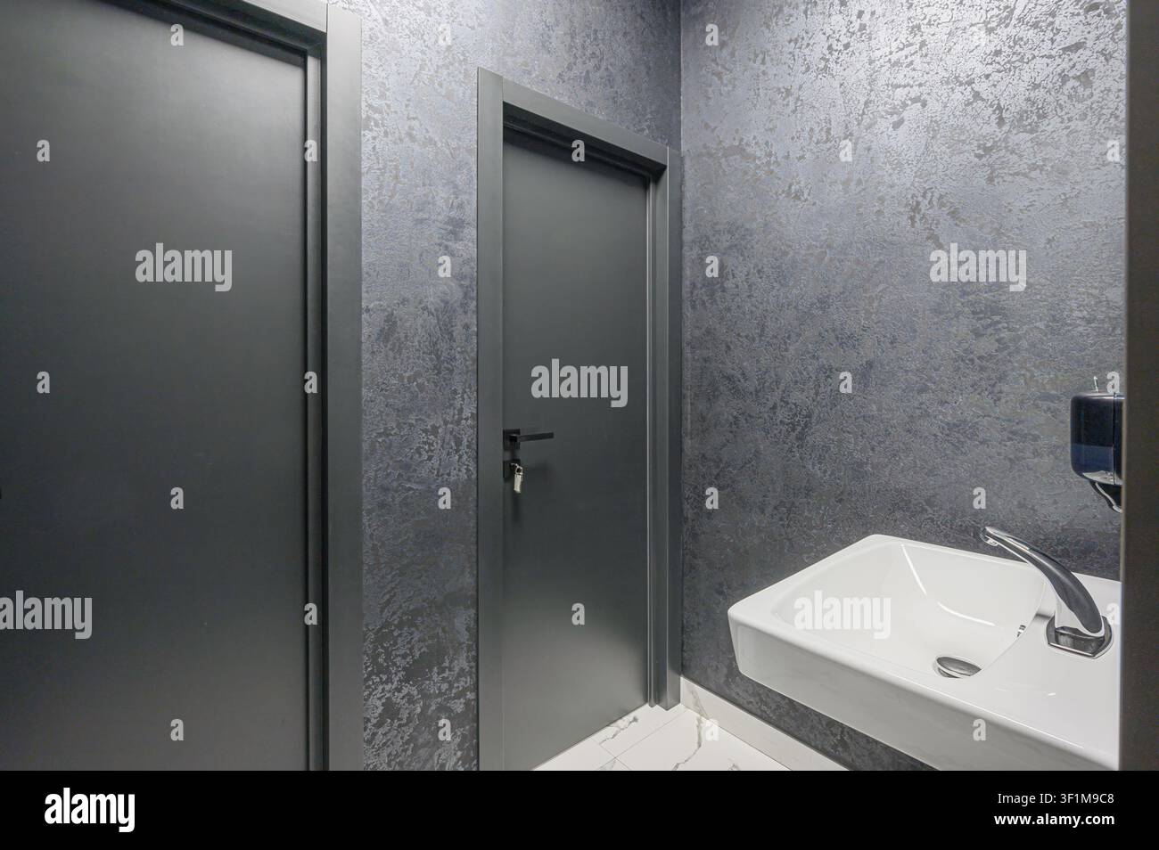 Moderner Waschraum mit zwei grauen Türen, strukturierten grauen Wänden und einem schlanken weißen Waschbecken mit silbernem Wasserhahn. Der Boden ist aus weißem Marmor mit grauen Adern Stockfoto