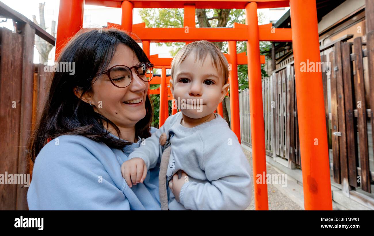 Ein freudiger Moment fängt eine Mutter ein, die warm lächelnd ihr Kind zwischen den lebhaften roten Torii-Toren des Horikoshi-Schreins in Osaka, Japan, betrachtet. Eine wunderschöne Mischung Stockfoto