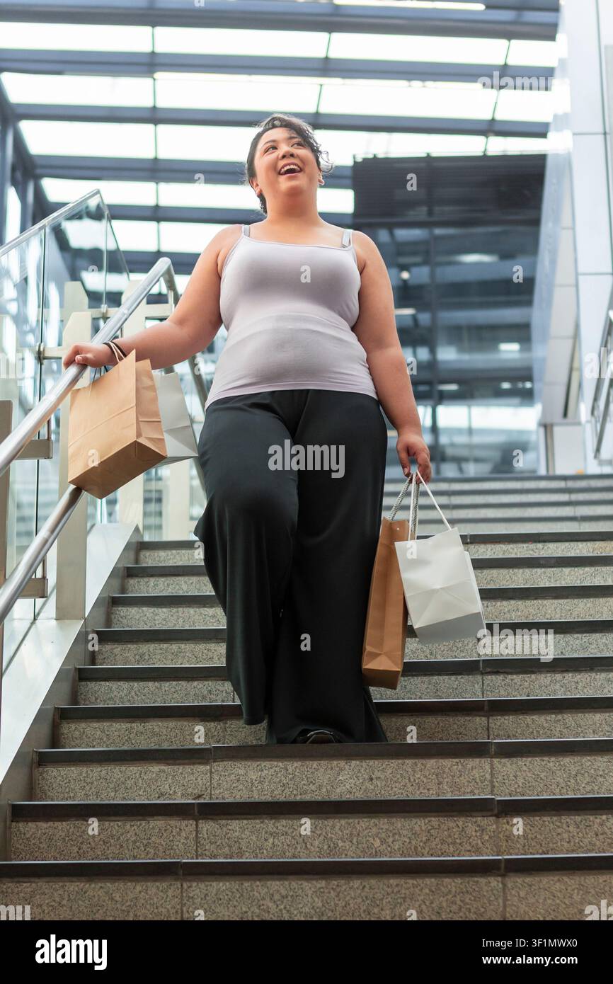 Lächelnde Frau trägt Papiereinkaufstüten, während sie auf Außentreppen eines modernen Stadt Einkaufszentrums hinuntergeht. Lässiges Outfit, helles Tageslicht, urbaner Lebensstil und Stockfoto