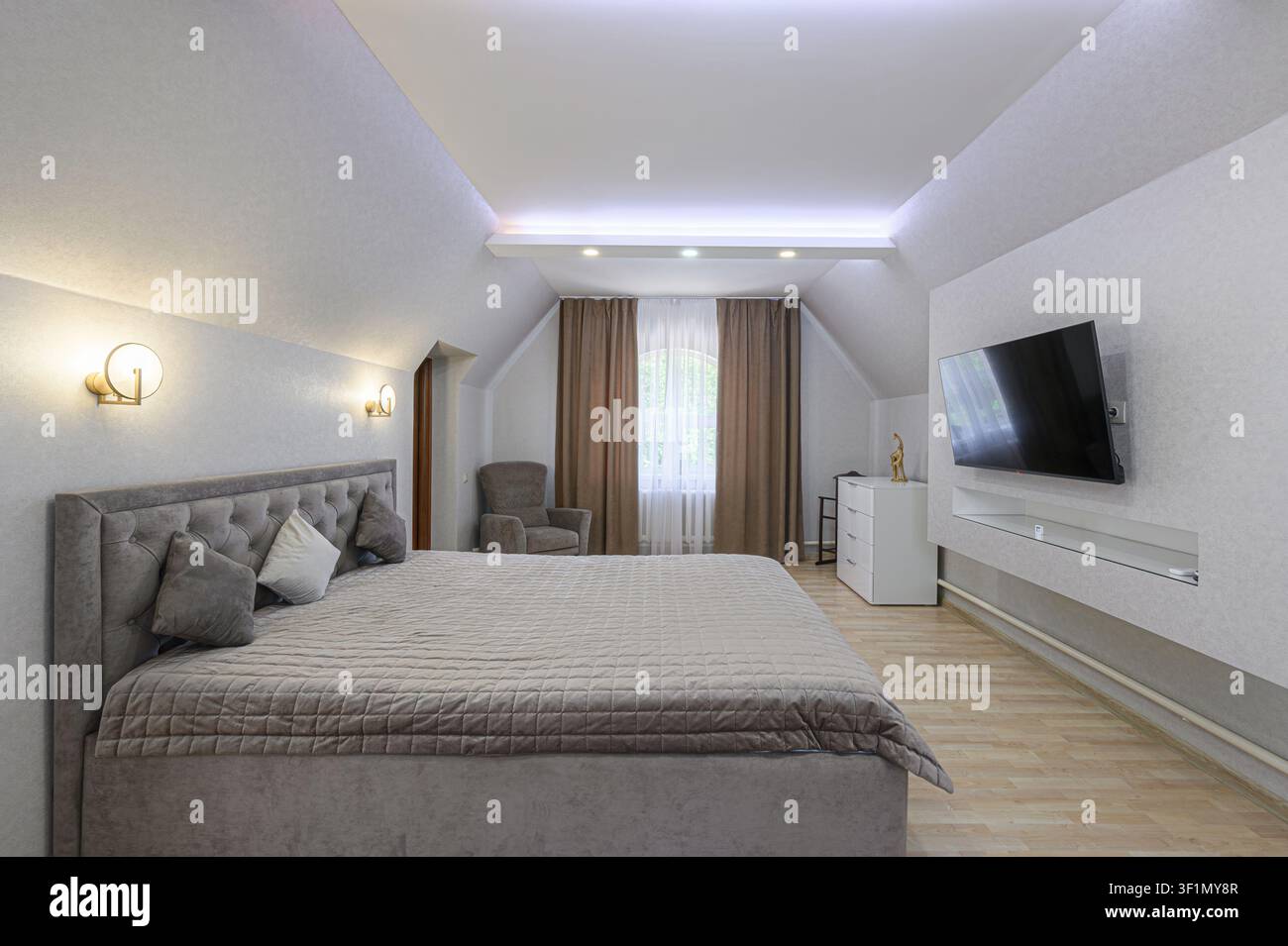 Gemütliches Dachgeschoss-Schlafzimmer mit großem Bett, Fernseher, Sessel und sanftem Licht. Der Raum wirkt ruhig und einladend Stockfoto