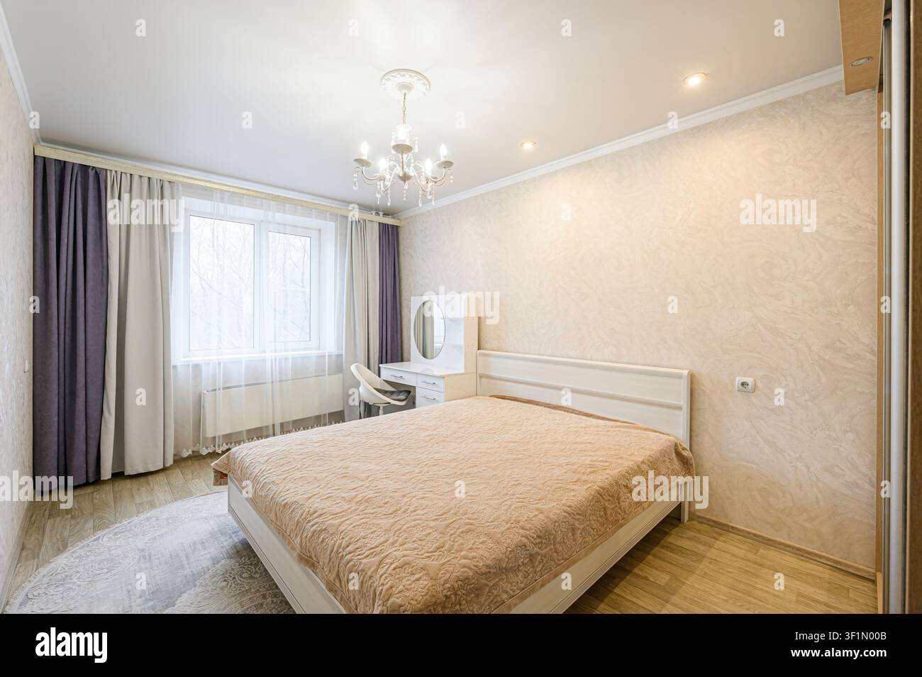 helles Schlafzimmer mit einem Bett, Schminktisch, Kronleuchter und Vorhängen. Der Raum ist ordentlich mit ruhiger Dekoration Stockfoto