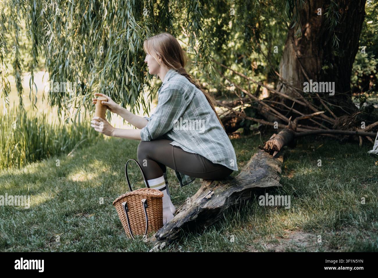 Frau sitzt auf einem Baumstamm in der Nähe einer Trauerweide und hält einen hölzernen Behälter, Outdoor-Wellness-Set, Tagesausflugsplanung, gesunde Snack-Zubereitung, Picknick-Zubehör Stockfoto