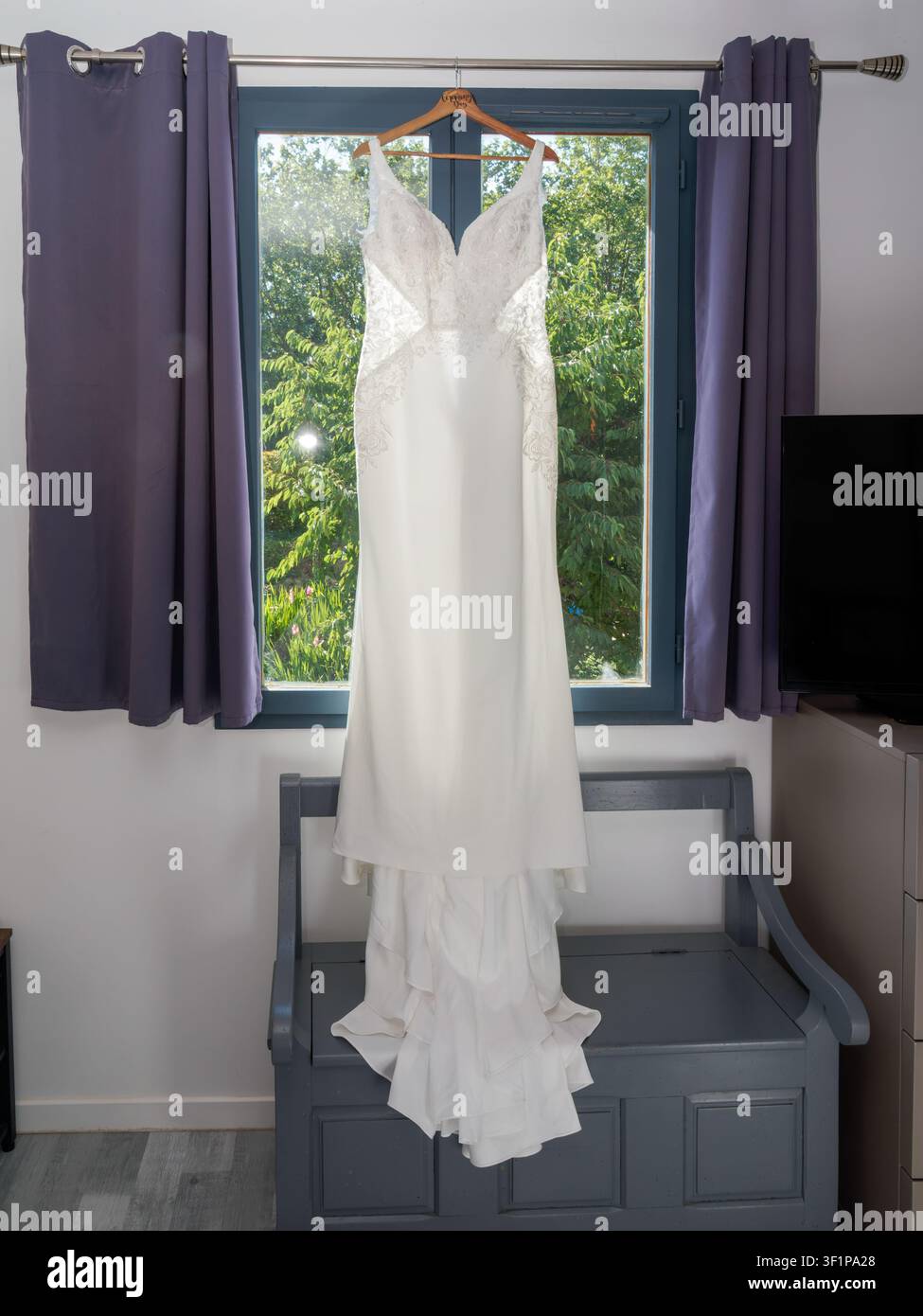 Elegantes weißes Spitzenhochzeitskleid hängt vor einem Fenster vor der Zeremonie Stockfoto