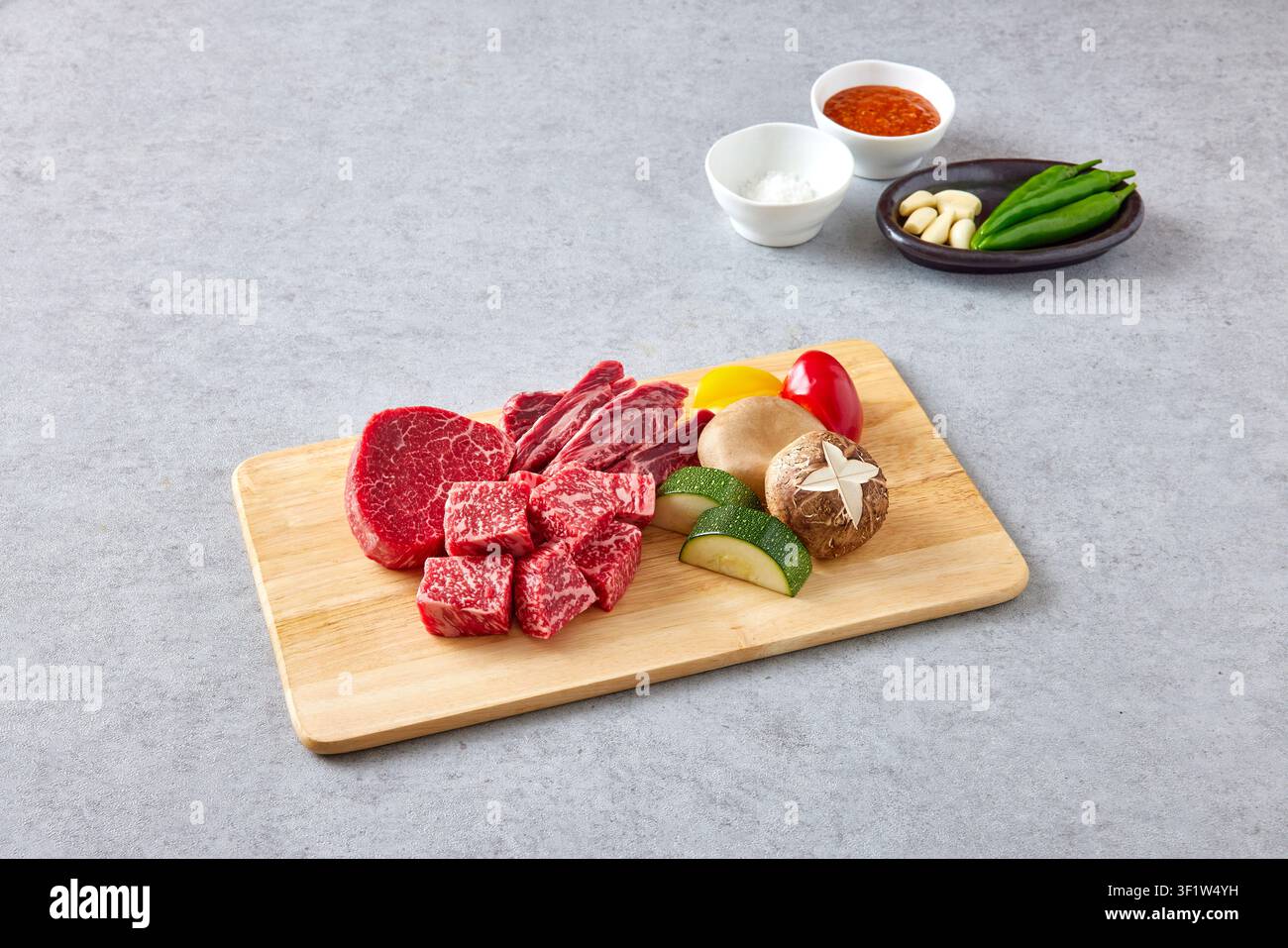 Premium koreanische Hanwoo-Wagyu-Rindfleischstücke mit starker Marmorierung, die auf einem Holzschneidebrett mit frischem Gemüse inklusive grüner Paprika und Pilz präsentiert werden Stockfoto